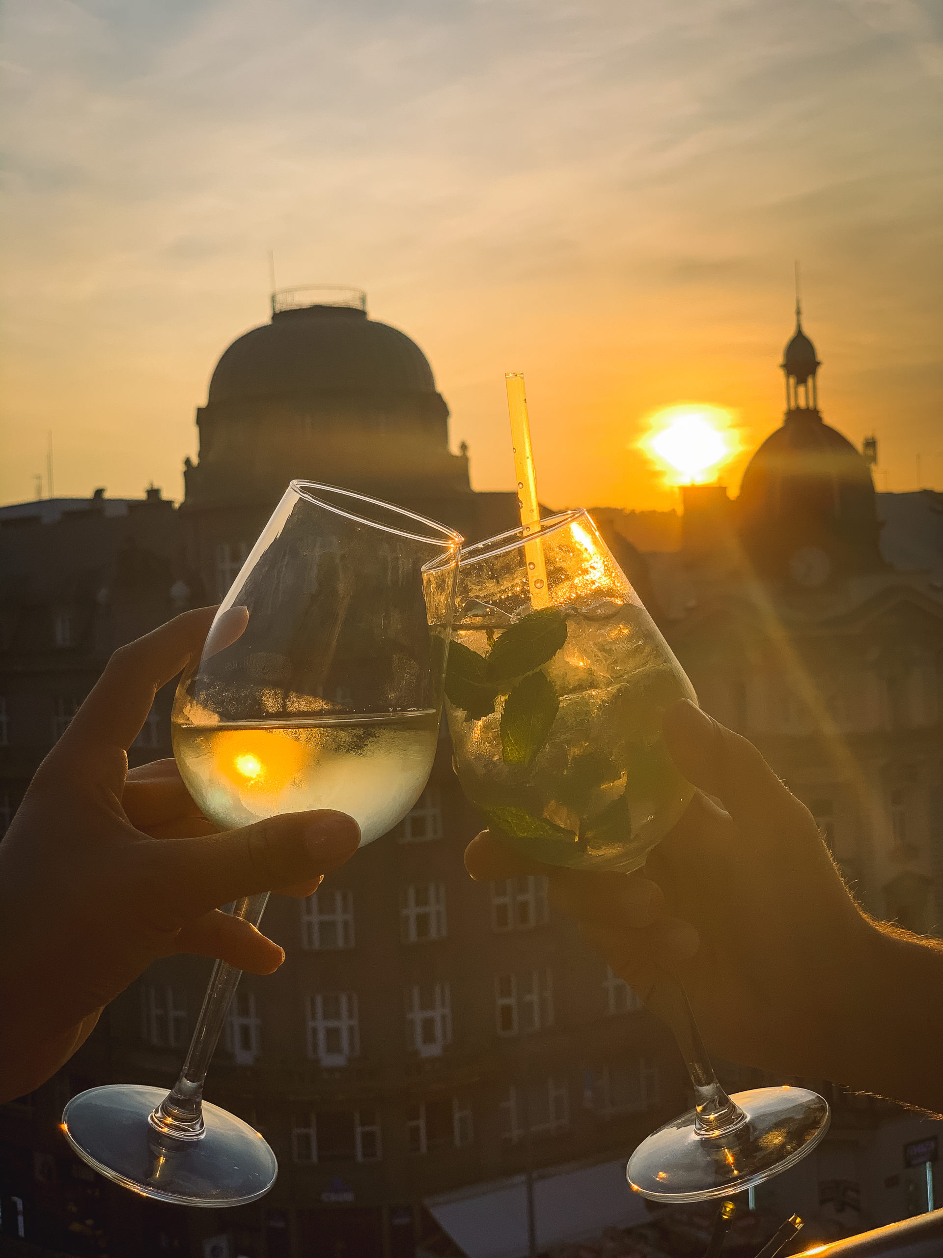 Two people cheering with two cocktails on a rooftop at sunset in Prague